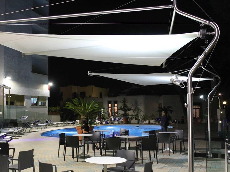 Vue nocturne d'une piscine extérieure éclairée avec tables, chaises et voiles d'ombrage.
