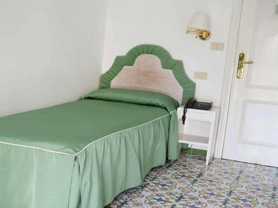 Jednolůžko s zeleným povlečením a květinovým kobercem v jednoduché místnosti.