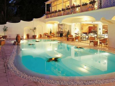 Večerní venkovní pohled na hotel s osvětleným bazénem a posezením.