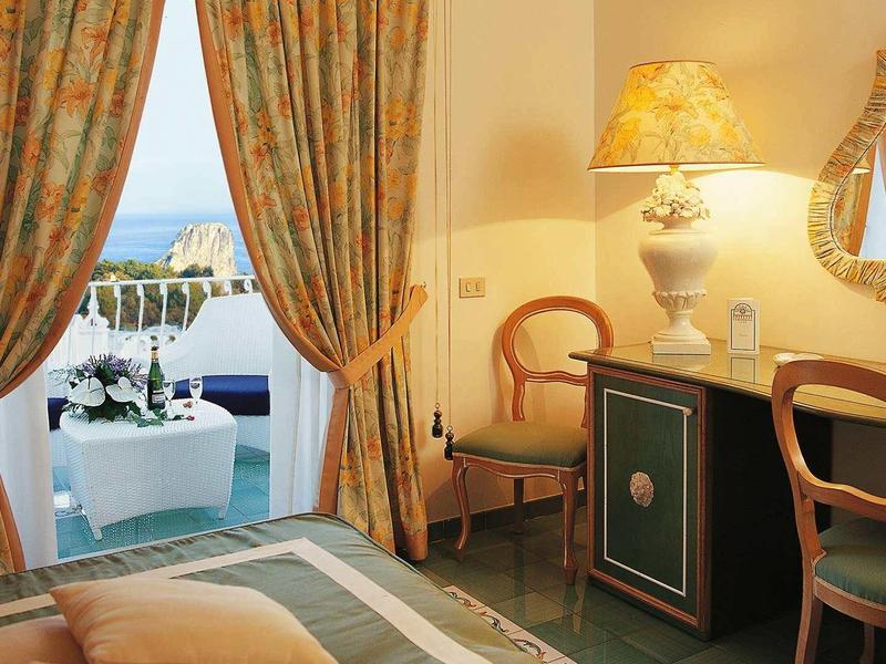Hotelový pokoj s výhledem na pobřeží, závěsy, psací stůl a židle vedle balkónu