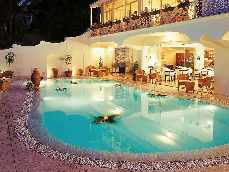 Večerní venkovní pohled na hotel s osvětleným bazénem a posezením.