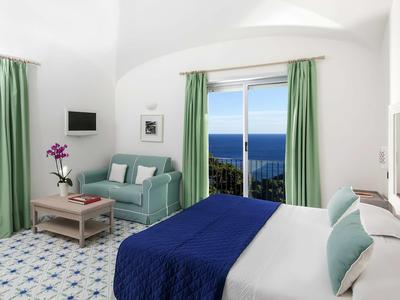 Chambre d'hôtel lumineuse avec lit double, couvre-lit bleu, canapé et vue sur la mer.