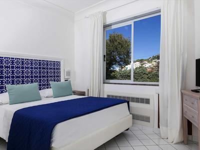 Chambre d'hôtel lumineuse avec lit double, couverture bleue et vue par la fenêtre.