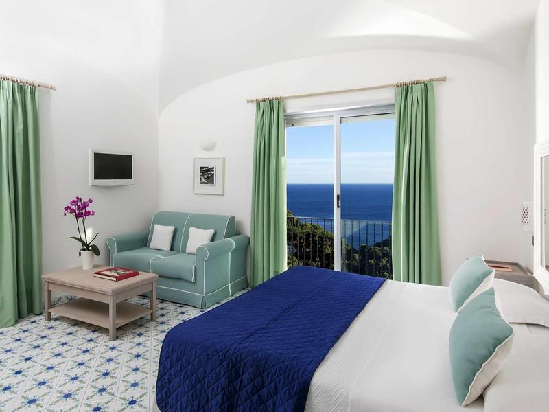 Chambre d'hôtel lumineuse avec vue sur la mer, rideaux bleus, lit double et canapé.