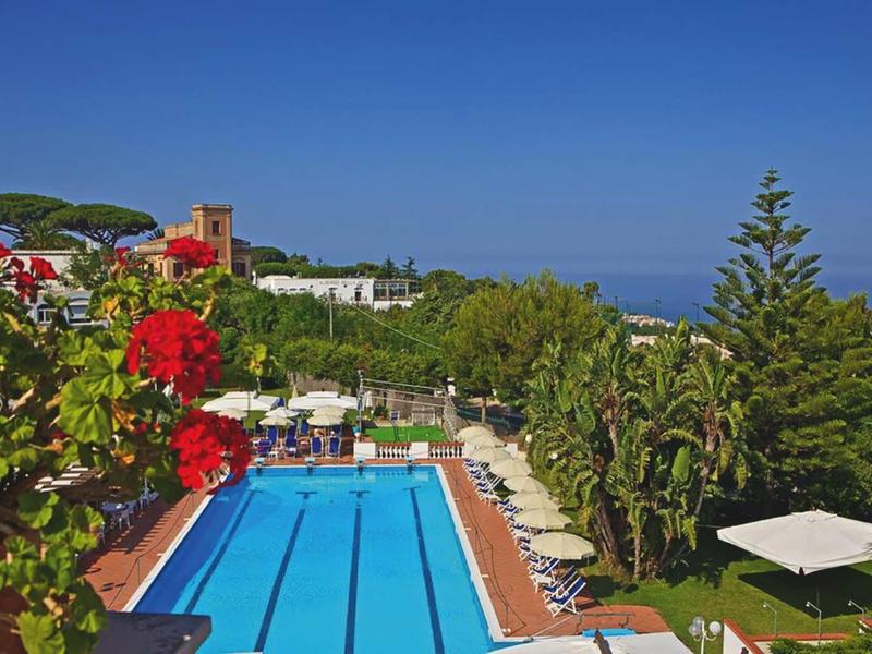 Outdoor pool with lounge chairs surrounded by plants and buildings under clear blue sky.