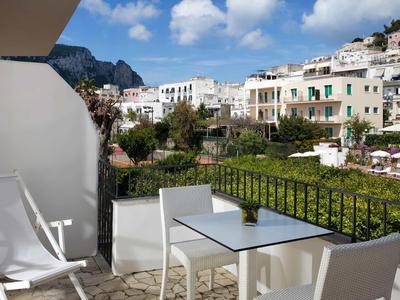 Balkon s bílými židlemi a stolem s výhledem na zelené zahrady a bílé budovy pod modrou oblohou.