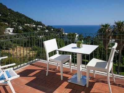 Balkon s bílým stolem a židlemi s výhledem na moře a zelené kopce.