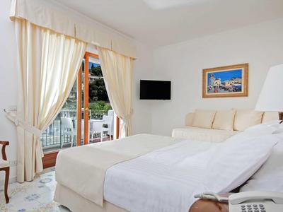 Lichte hotelkamer met tweepersoonsbed, gordijnraam, tv en stoel.