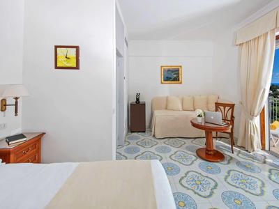 Lichte hotelkamer met terras, bank, fauteuil en uitzicht op de blauwe zee bij zonnig weer.