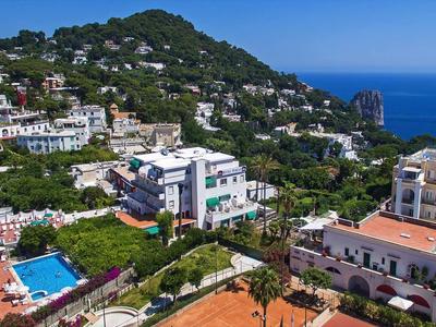 Pohled na přímořské letovisko s několika hotely, bazénem a čistým výhledem na moře za jasné oblohy.