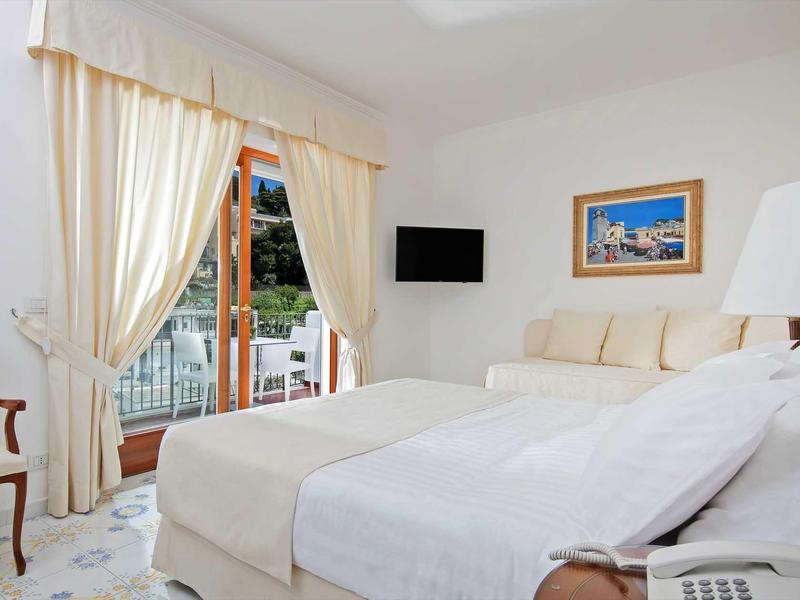 Lichte hotelkamer met tweepersoonsbed, gordijnraam, tv en stoel.