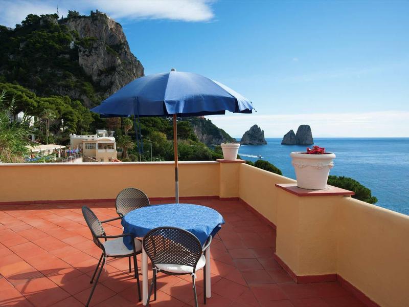 Terras met blauwe tafel, stoelen en parasol met uitzicht op zee en rotsformaties.
