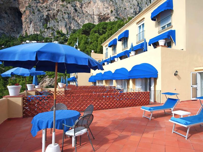 Terras met blauwe parasols, stoelen en ligbedden voor een hotelgebouw met kliffen op de achtergrond.