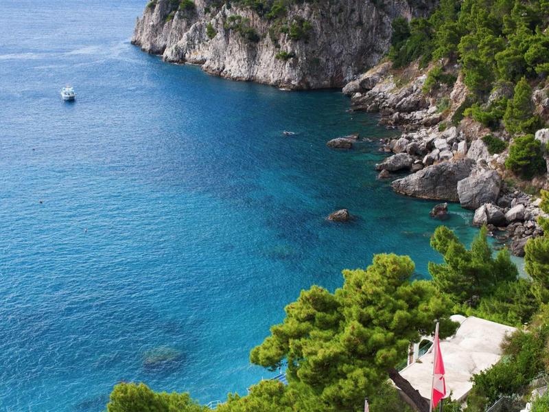 Helder blauw water langs steile kust met groene bomen en een kleine boot in de verte.