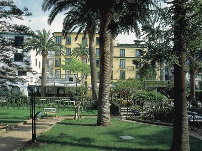 Grüner Park mit Palmen, Bänken und einem gelben Gebäude im Hintergrund bei Tageslicht.