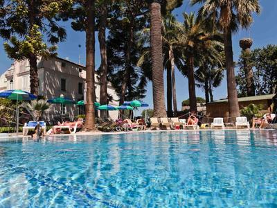 Großer blauer Pool mit Liegestühlen, Sonnenschirmen und hohen Palmen unter blauem Himmel.