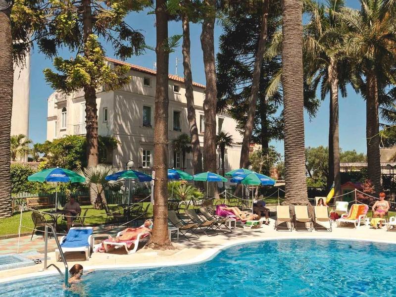 Ein blauer Pool vor einem Haus, umgeben von großen Bäumen und Sonnenschirmen bei sonnigem Himmel.