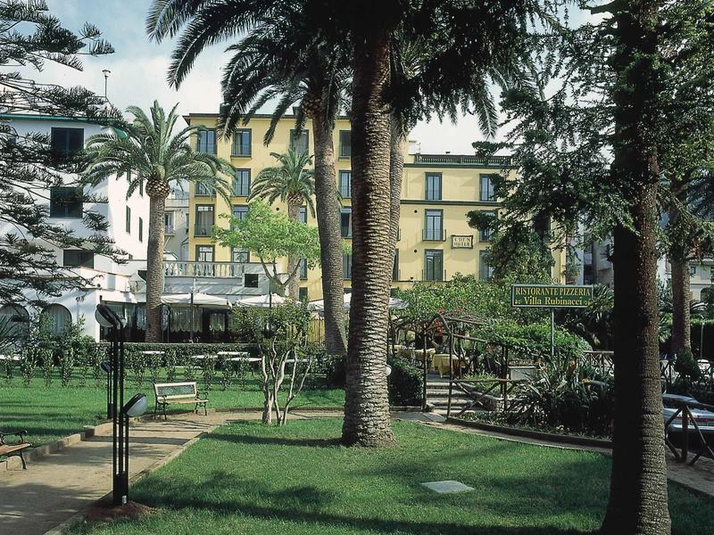 Grüner Park mit Palmen, Bänken und einem gelben Gebäude im Hintergrund bei Tageslicht.