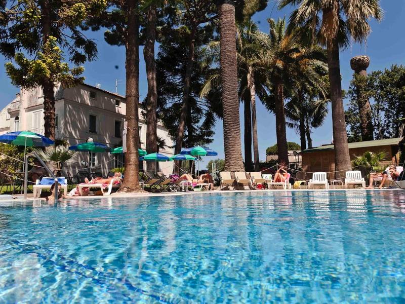 Großer blauer Pool mit Liegestühlen, Sonnenschirmen und hohen Palmen unter blauem Himmel.