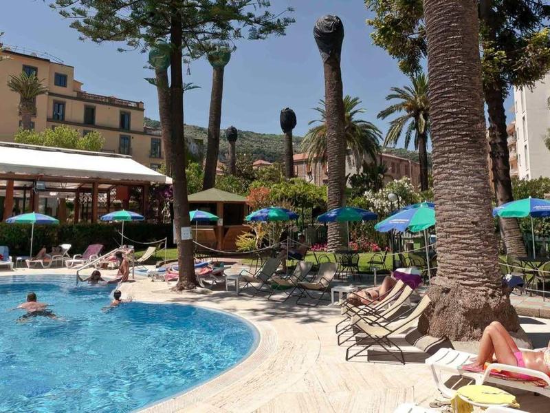 Ein Außenpool mit Menschen, umgeben von Palmen, Sonnenschirmen und Hotelgebäuden bei Sonnenschein.