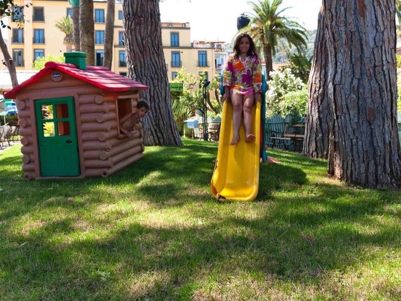 Kind in gelbem Kleid schaukelt auf einem Spielplatz mit Spielhäusern und Bäumen im Hintergrund.