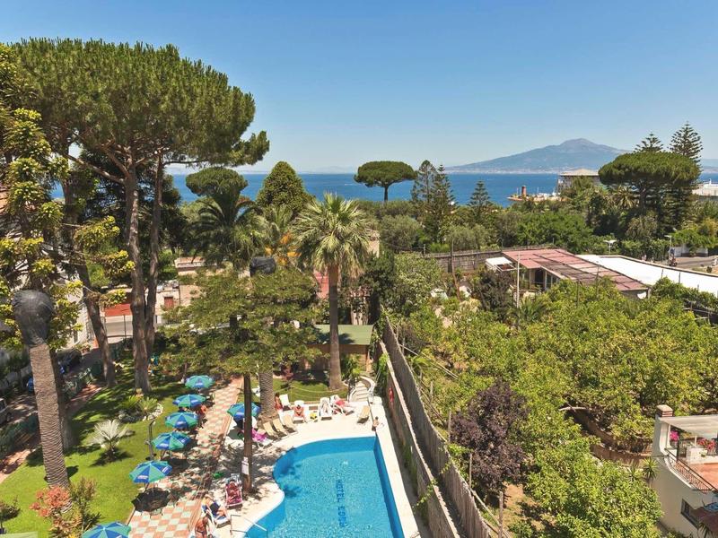 Blick auf einen Pool mit blauen Liegestühlen, Palmen und dahinter das Meer und Berge.