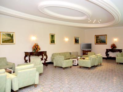 Helles Hotelwartezimmer mit grünen Sesseln, Couchtischen und Gemälden an den Wänden.