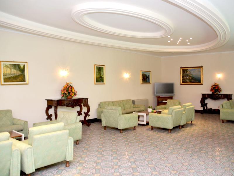 Jasny poczekalnia z zielonymi fotelami, małymi stolikami, obrazami i telewizorem.