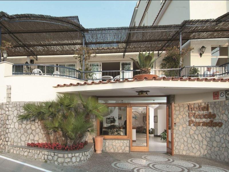 Wejście do hotelu z kamiennym murem, roślinami i zadaszonym balkonem na górze.