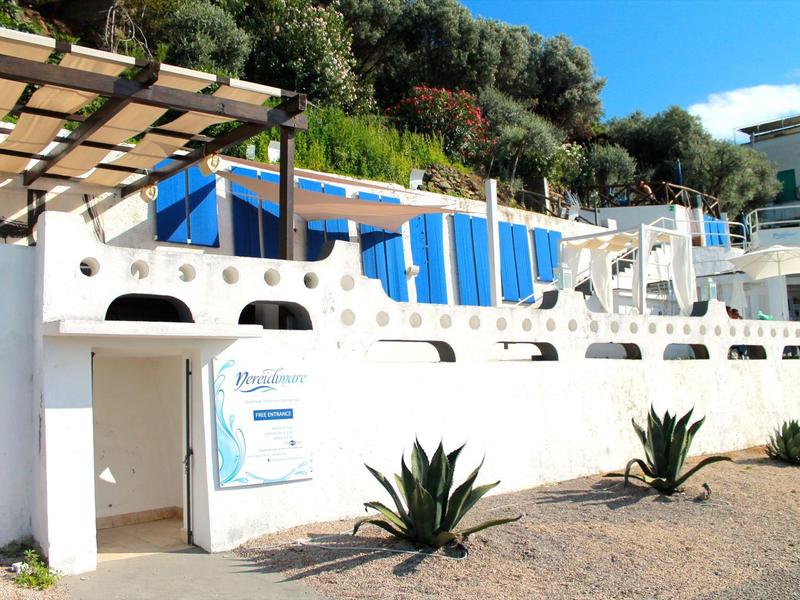 Biały budynek z łukowatymi oknami i niebieskim markizą, otoczony roślinami i drzewami