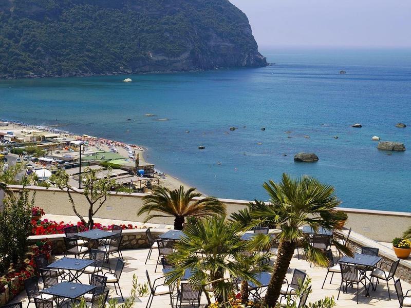 View of a coastal bay with blue water, tables, chairs, and palm trees on the beach.