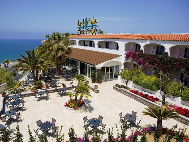 Hotel terrace with tables, chairs, and palm trees overlooking the sea under clear sky.