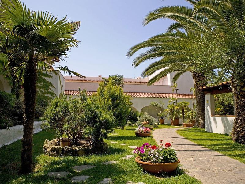 Green garden with palm trees and colorful flowers beside a paved path under blue sky.