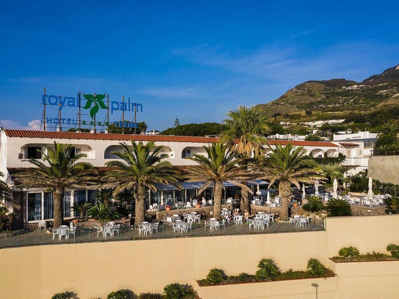 Hotel restaurant with terraces, palm trees, and mountain backdrop under a blue sky.