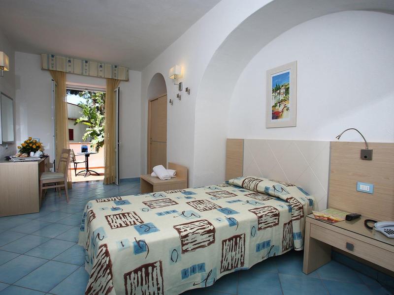 Bright hotel room with single bed, desk, and balcony access overlooking garden.