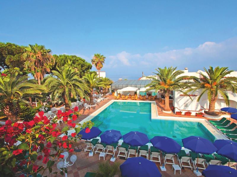 View of a hotel pool with lounge chairs and palm trees under a blue sky.