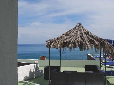 Strand mit blauem Himmel, Meer, Palmdach-Sonnenschirm und schwarzer Sitzgruppe auf Terrasse.