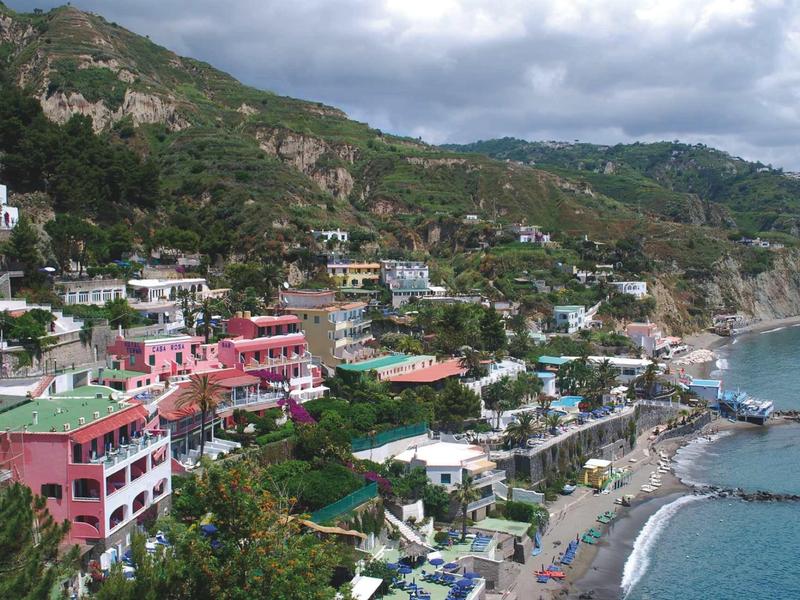 Küstenstadt mit bunten Häusern, grünen Hügeln und grauem Strand unter bewölktem Himmel.