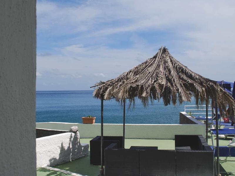 Strand mit blauem Himmel, Meer, Palmdach-Sonnenschirm und schwarzer Sitzgruppe auf Terrasse.