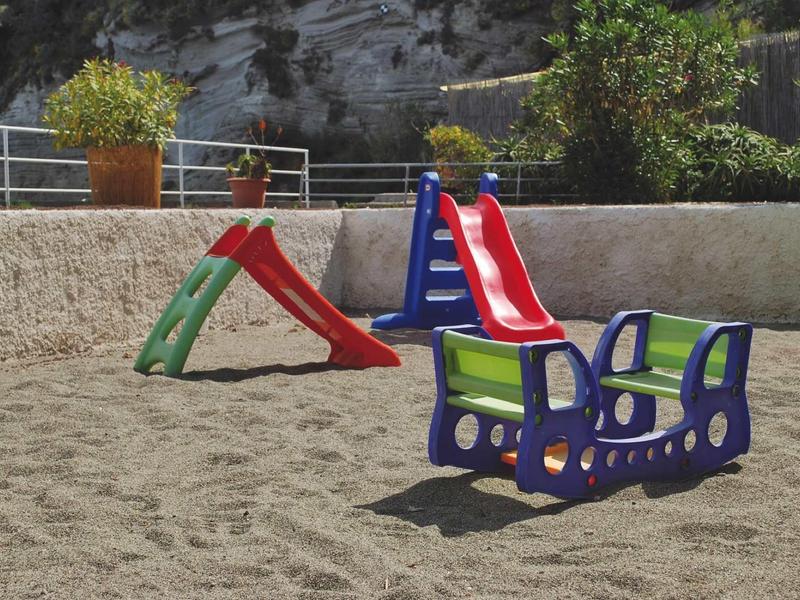 Kinderspielplatz mit bunten Rutschen und Klettergeräten auf sandigem Boden und Felswand im Hintergrund.