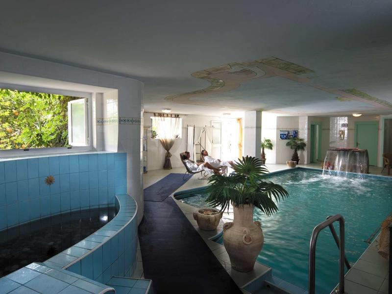 Innenraum mit großem Schwimmbecken, blauen Fliesen, Pflanzen und natürlichem Licht durch Fenster.