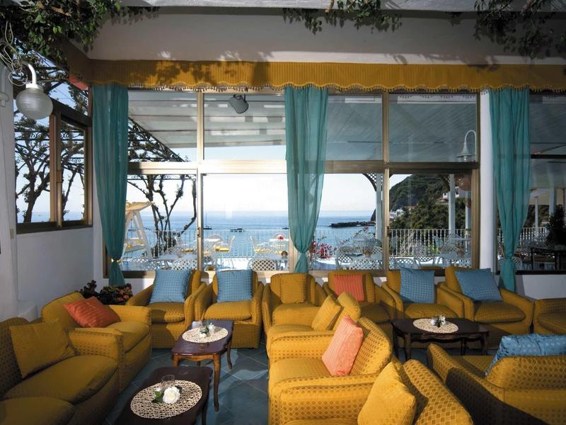 Gemütliche Lounge mit gelben Sofas, grünen Vorhängen und Blick aufs Meer bei Sonnenuntergang.