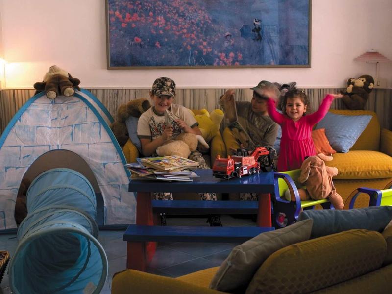Kinder spielen in einem gemütlichen Raum mit gelber Couch, blauem Spielzelt und buntem Teppich.
