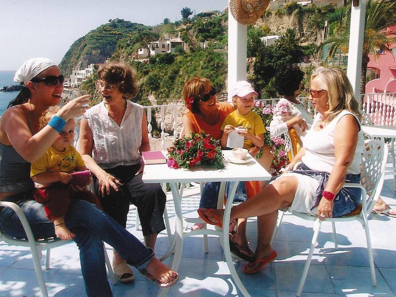 Vier Frauen sitzen entspannt an weißem Tisch mit blauen Stühlen, Meer und Häuser im Hintergrund.