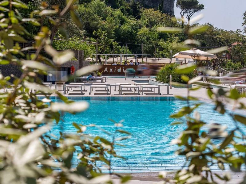 Area piscina con lettini accanto a un hotel, circondata da alberi verdi e ombrelloni.