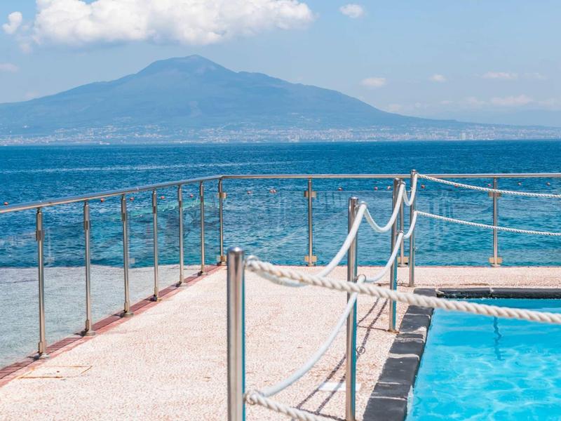 Vista di una piscina sul mare con montagne sullo sfondo in una giornata di sole.