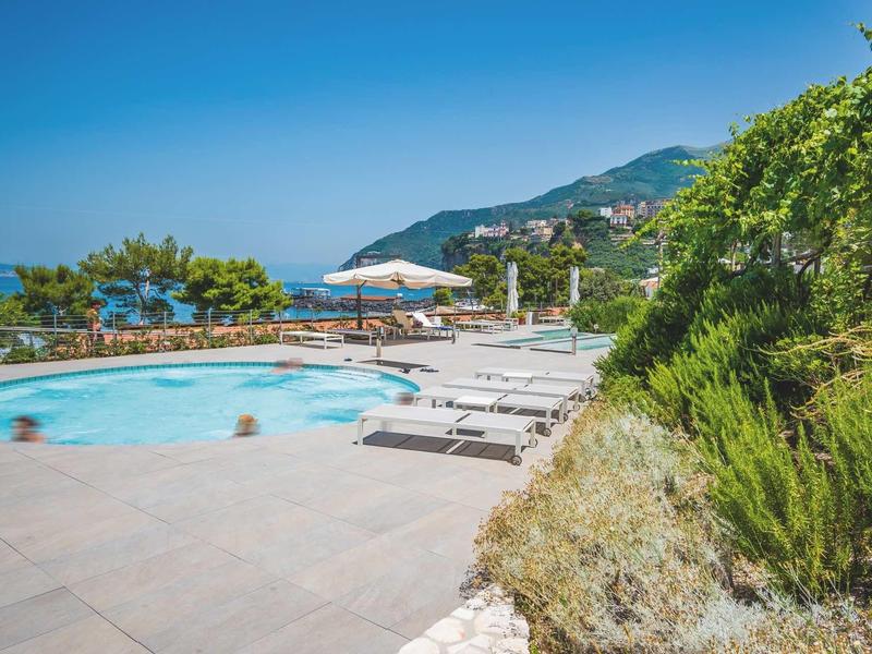 Piscina rotonda con lettini e ombrelloni, mare e montagne sullo sfondo.
