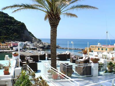 Výhled na terasu s posezením a palmou, v pozadí hora a moře pod jasnou oblohou.