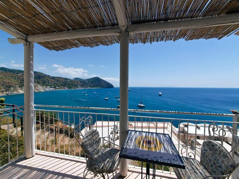Balkón se stolem a židlemi s výhledem na modré moře a pobřeží pod stříškou z bambusových rohoží.