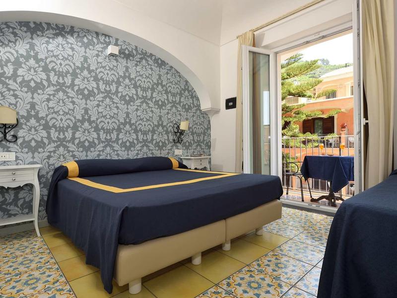 Bright hotel room with double bed, floral wallpaper, and terrace access overlooking garden.